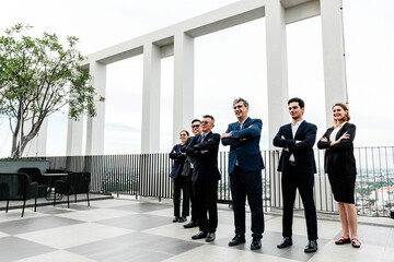 Confident business team in formal attire standing with arms crossed on a modern rooftop terrace, with city skyline and geometric architecture in the background.
