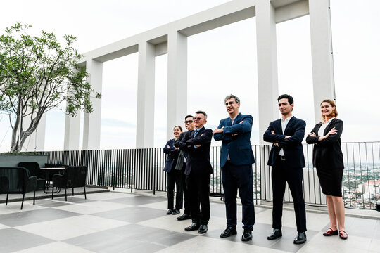 Confident business team in formal attire standing with arms crossed on a modern rooftop terrace, with city skyline and geometric architecture in the background. - Powered by Adobe