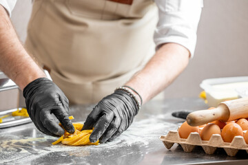 Chef preparing fresh homemade pasta on floured surface with eggs, rolling pin, and pasta machine. Hands in black gloves shaping yellow tagliatelle. Culinary process in kitchen