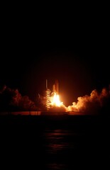 Dramatic vertical shot of a nighttime rocket launch, flames illuminating the smoke and clouds against the dark sky and water