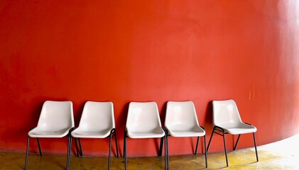 Five white plastic chairs lined up in front of a bold curved red wall, evoking simplicity, waiting, and modern design.