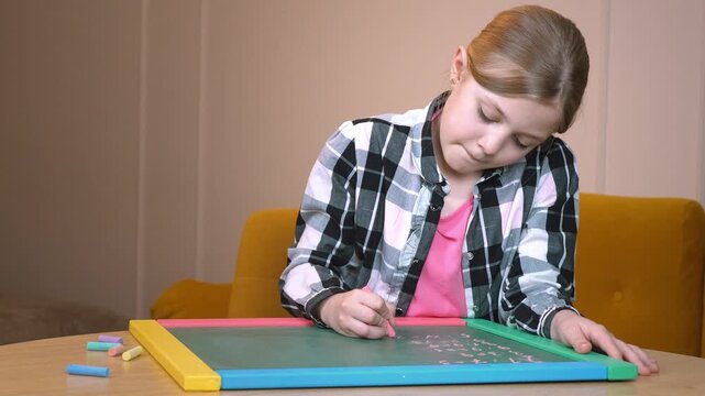 Primary school student practicing drawing on a small blackboard placed on a round wooden table near a mustard-colored sofa in a cozy living room in an apartment
