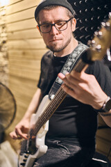 Obraz premium Young adult man with short hair, glasses plays bass guitar in cozy studio with wooden panels. Intense focus on music, warm lighting enhances creative atmosphere