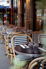 Tables are set at an empty outdoor Parisian cafe. The focus is on a round bistro table with classic black and white wicker chairs.