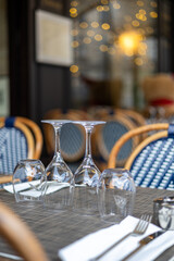 Close-up of a set table at an outdoor Parisian cafe, focusing on wine glasses. Classic blue and white wicker chairs and bokeh lights are in the background.
