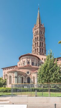 The cobbled parvis of the Basilica of Saint Sernin in Toulouse, France, timelapse hyperlapse. Largest romanesque church in Europe with brick bell tower, arched doors, green trees and blue sky