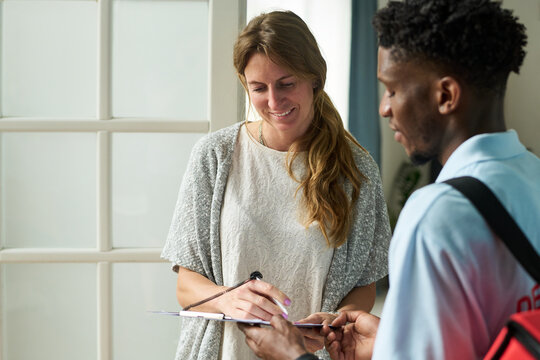 Woman smiling while signing document on clipboard held by Black young man standing indoors, both engaging in friendly interaction during delivery or service - Powered by Adobe