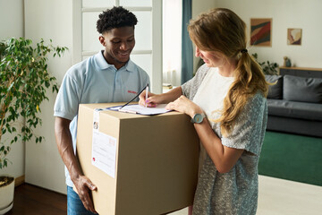 Woman signing delivery form while Black young man holding large cardboard box, both standing in...