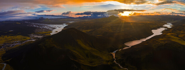 Aerial view of Iceland's rivers and lakes amid moss covered hills, with a glacier in the distance and golden sunlight breaking through clouds.
