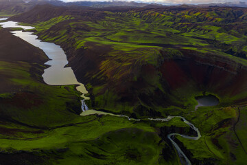 Aerial view of Iceland's green terrain with volcanic soil, a crater with a lake, a winding river, a waterfall, and an elongated lake on the left.