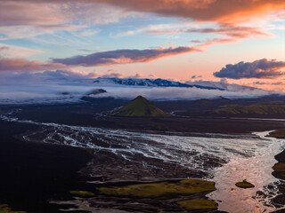 A green volcanic hill rises from dark plains, surrounded by glacial rivers. Snow capped mountains and clouds enhance the sunset lit Icelandic landscape.