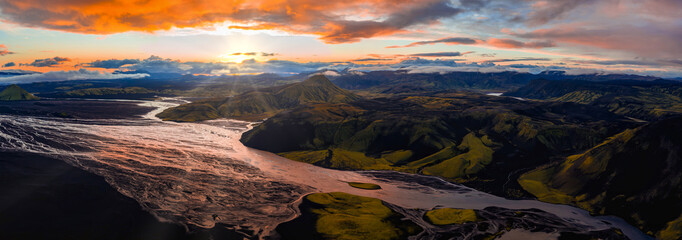 A glacial river winds through volcanic terrain in Iceland, surrounded by green hills and rugged mountains under a golden sunset with dramatic clouds.