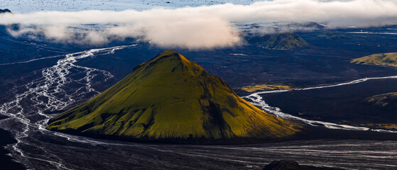 Aerial view of a moss covered Maelifell volcanic cone surrounded by dark plains, with reflective glacial rivers cutting through the terrain under low clouds in Iceland.