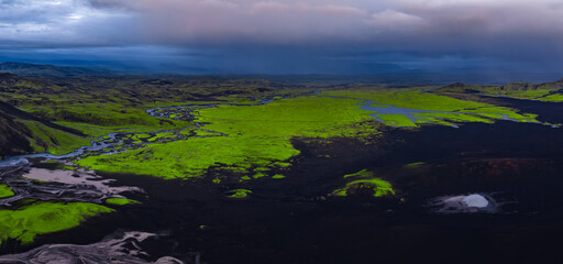 Aerial view of Iceland's moss covered plains, dark volcanic terrain, meandering rivers, and a cloudy sky with a small patch of snow or ice visible.
