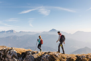 Two hikers, a man and a woman, walking over a grassy slope with a scenic view of misty mountains...