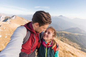 Fototapeta premium Young couple smiles at each other, embracing the stunning mountain view during their hiking adventure in nature.