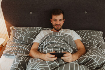 Relaxed young man smiling while scrolling on phone in bed