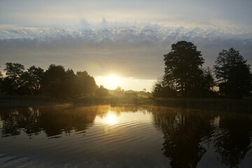 Beautiful moments of sunrise. Shot from the surface of the lake, the shore and trees against the beautiful sky. Morning fog. Reflection on the surface of the water.
