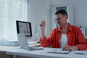 Naklejka premium Focused mature businessman in a red shirt analyzing financial charts and working on a laptop while reviewing documents in a modern office space filled with natural light