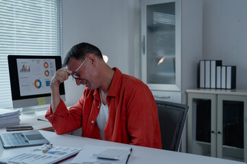 Businessman massaging his temples while dealing with a headache, working on a laptop and analyzing financial charts at his desk in a corporate office, feeling the weight of stress and pressure