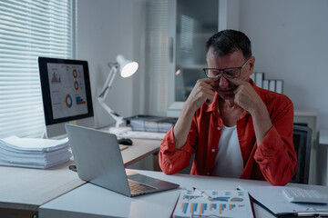Businessman sitting at his desk rubbing his eyes, showing tiredness and fatigue, while working with a laptop and financial reports in a modern office