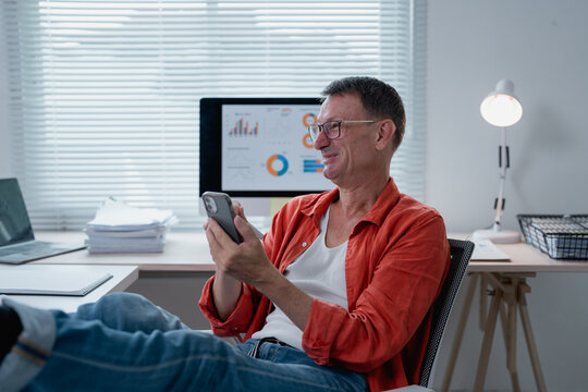 Senior businessman relaxing with feet up on desk, using smartphone and smiling in modern office, while computer displays graphs and charts in the background - Powered by Adobe
