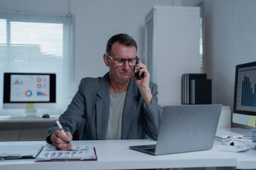 Obraz premium Worried mature businessman talking on phone and reviewing negative financial charts, feeling stressed and frustrated about bad news and financial losses in office