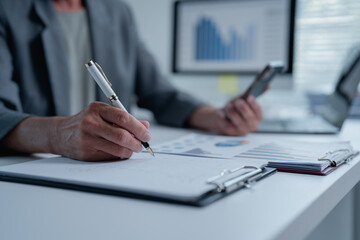 Businesswoman is using smartphone and analyzing financial charts while taking notes with a pen on clipboard during a working day in the office