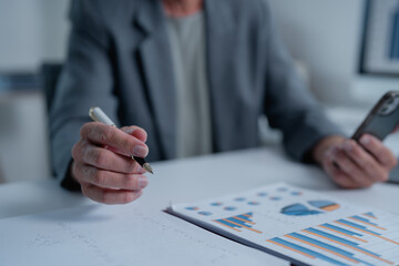 Senior businessman holding a pen and using a smartphone, analyzing financial charts and taking notes while seated at an office desk, focusing on strategy and market growth