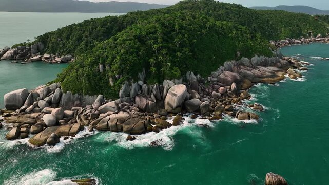 Aerial view of a rugged coastline where the turquoise ocean meets a dense green forest, with scattered rocks adding texture to the landscape, Paraty, State of Rio de Janeiro, Brazil.
