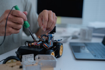 Engineer is soldering electronic components of a robot car prototype in a laboratory using a soldering iron and tin wire, with a laptop and documents in the background
