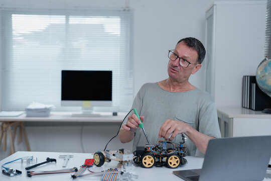 Electronics engineer working on a programmable robotics toy car using a soldering iron and various components on a white table in his laboratory, next to a desktop computer and a laptop