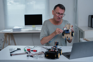 Engineer connecting cables to a robot chassis while following instructions on a laptop, in his home office equipped with a computer and tools for electronics and robotics projects
