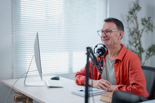 Radio host speaking with microphone and smiling during podcast recording in his home studio, he's sitting at his desk with a notebook and a computer - Powered by Adobe