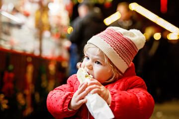 Little baby girl, cute child eating bananas covered with chocolate, marshmellows and colorful sprinkles near sweet stand with gingerbread and nuts. Happy toddler on Christmas market in Germany.