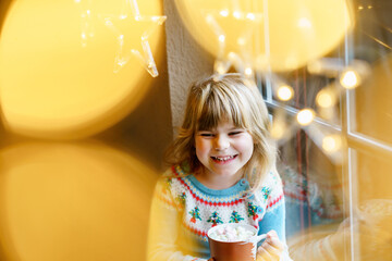 Little preschool girl holding cup with hot chocolate with marhsmallows. Happy child drinking sweet...