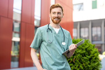 Smiling man carrying laptop in hands near hospital wall
