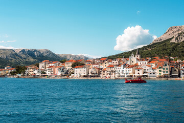 Obraz premium Coastal town with red roof houses, mountains in background and blue sea in front. Baska town, Krk island, Croatia