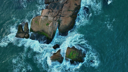 Aerial view of rugged, moss-covered rocks meet the swirling turquoise sea, creating a dynamic contrast of textures and tones, Paraty, State of Rio de Janeiro, Brazil.