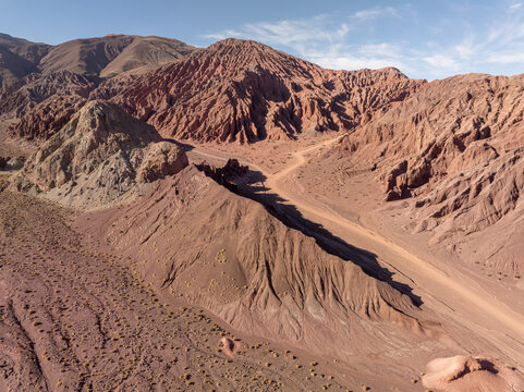 Aerial view of a sinuous dirt road carving through the ochre and crimson landscape of Valle del Arcoiris, a tapestry of geological wonder, Valle del Arcoiris, San Pedro de Atacama, Chile.