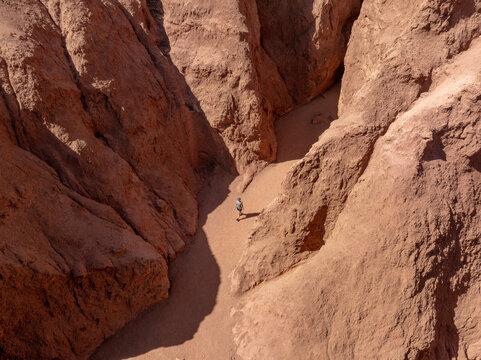 Aerial view of a lone figure walking through the sun-baked, ochre-red canyon of Valle del Arcoiris, the shadows deepening the textures of the arid landscape, San Pedro de Atacama, Chile.