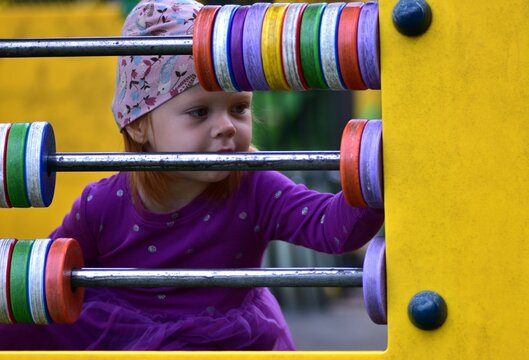 Close-up of toddler girl in purple dress and hat playing with colorful abacus toy at playground. Child moving beads while exploring fun learning activity outdoors.