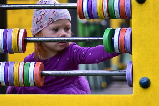 Close-up of toddler girl in purple dress and hat playing with colorful abacus toy at playground. Child moving beads while exploring fun learning activity outdoors.