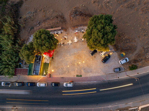 Aerial view of a cozy gathering spot under trees, illuminated by warm lights, next to a curving road with blurred vehicle light trails, Chania, Chania, Greece.