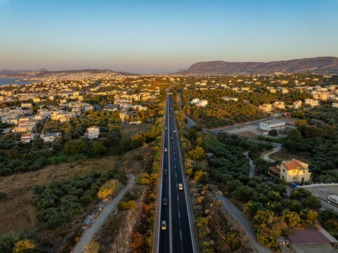 Aerial view of a long road cutting through a landscape dotted with houses and green trees under the soft light of the setting sun, Chania, Chania, Greece.