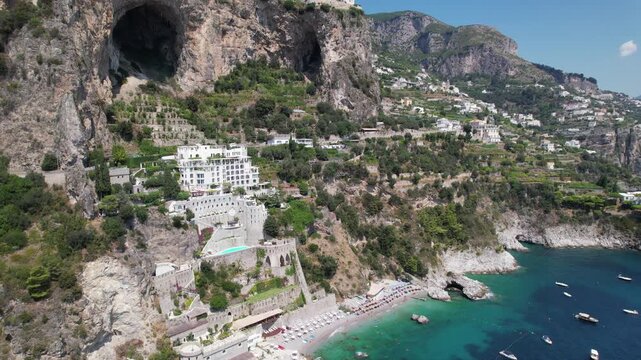 Smooth aerial pan of Conca dei Marini with dramatic cliffs, massive coastal cave, luxury villas, terraced gardens, and turquoise beach on the Amalfi Coast.