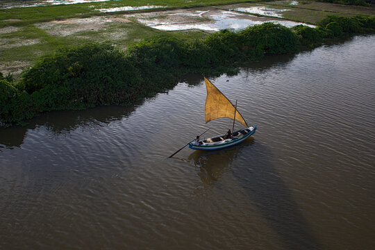 Yanam, India - 03 October 2010: Aerial view of a small boat with a striking yellow sail glides through the tranquil, murky river waters, bordered by vibrant green vegetation and rice paddies.