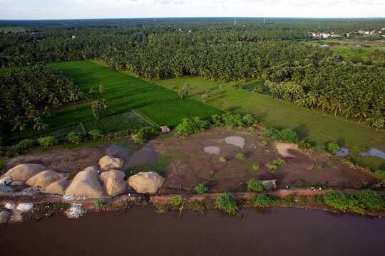 Yanam, India - 03 October 2010: Aerial view of the river's edge where piles of sand meet lush green fields, transitioning into a dense forest under a hazy sky.