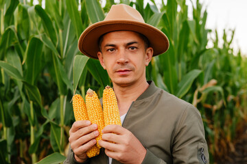 A focused portrait of a male farmer with a hat and jacket, holding three ripe corn cobs and looking...