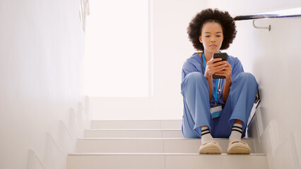 Female Nurse Wearing Scrubs Browsing On Mobile Phone Sitting On Stairs In Hospital